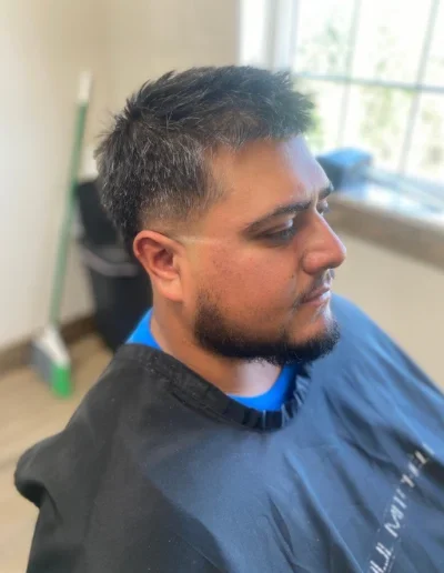Side profile of a man with a short beard wearing a barber cape during a haircut in a bright salon with a window in the background